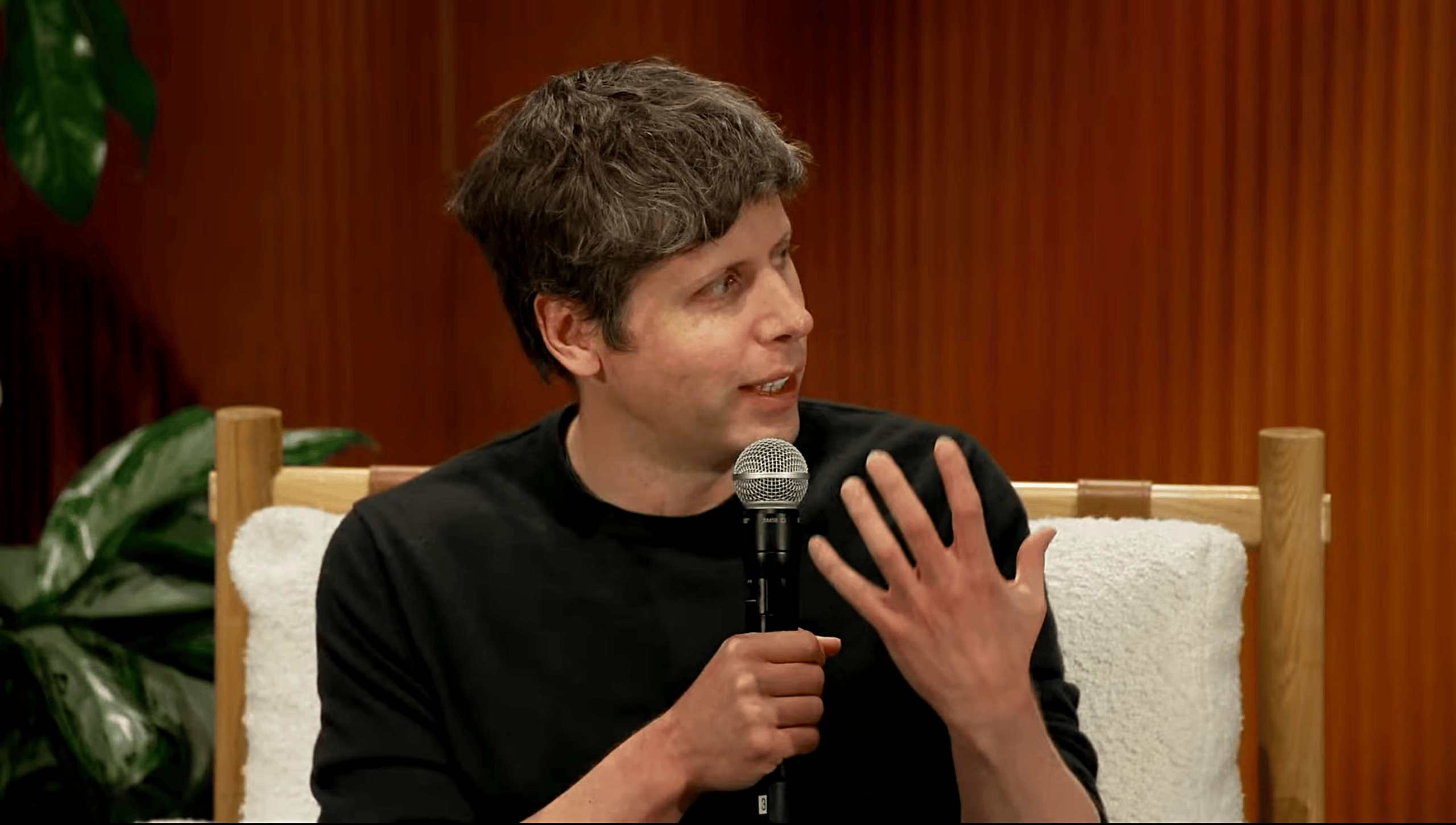 Sam Altman Confirms GPT-5.2X Level Intelligence Will Cost 100x Less by 2027 1 Sam Altman holding a microphone in a wood-paneled room, gesturing expressively. Green plant in the background, relaxed setting.
