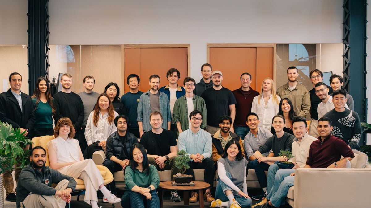 Goodfire Raises $150M Series B at $1.25B Valuation to Advance AI Interpretability 1 A group portrait of approximately thirty people, likely a professional team, posing together in a bright, modern office space. The group is arranged in three tiers: some are sitting on the floor or on sofas in the front, while others stand in two rows behind them. The individuals are dressed in casual and smart-casual attire, such as sweaters, button-down shirts, and t-shirts. In the center foreground, a small bonsai tree sits on a round wooden coffee table. The background features light-colored walls, large windows, and wooden door frames, creating a clean and collaborative atmosphere.