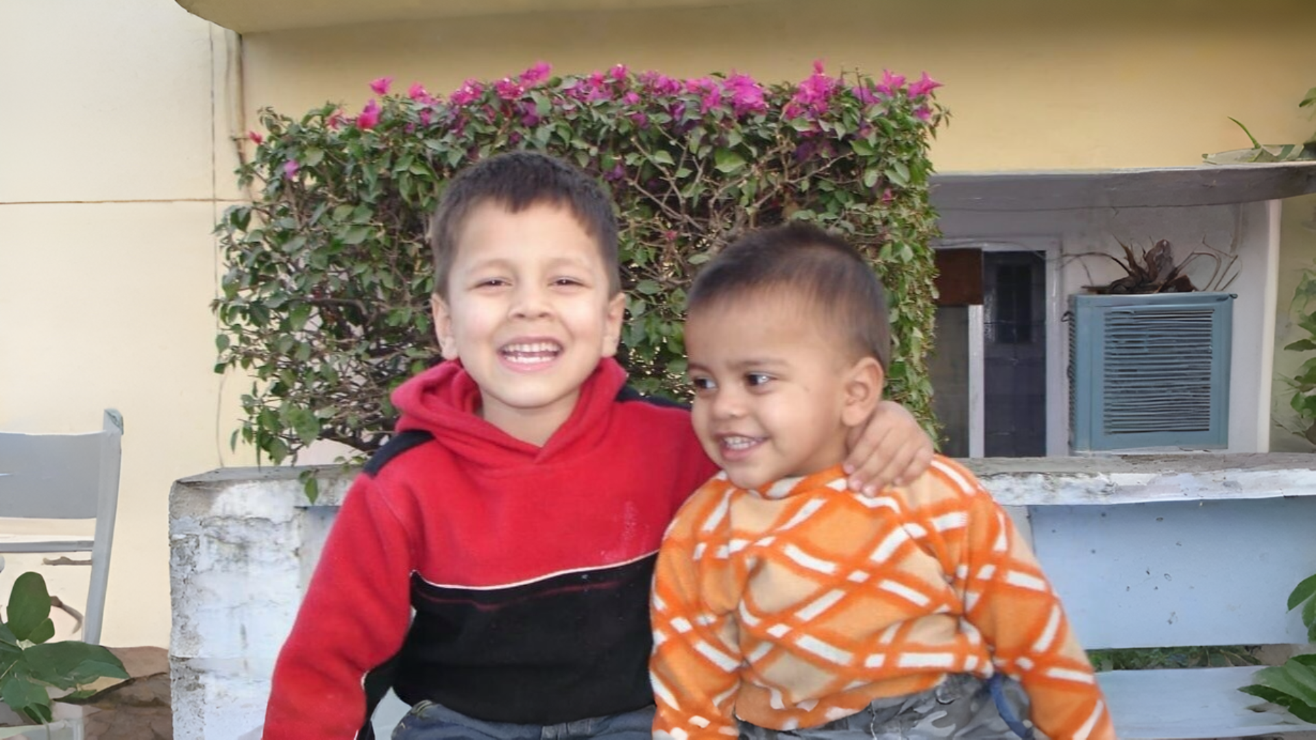 Two young boys sitting together outdoors. The older boy on the left, wearing a red and black hoodie, smiles broadly at the camera with his arm around the younger boy. The younger boy, wearing an orange and white patterned shirt, smiles while looking off to the side. They are sitting in front of a bush with pink flowers and a yellow wall.