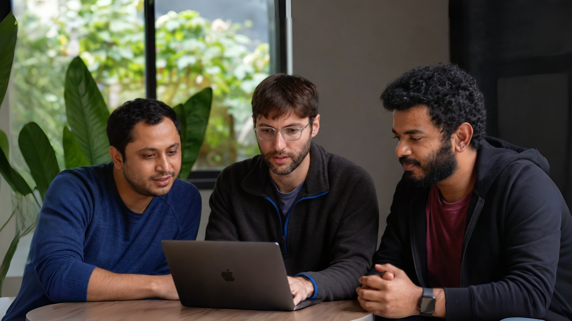 Three startup founders collaborating around a laptop in a modern office with natural daylight, soft background blur, and cinematic editorial photography style. The team sits at a wooden table discussing ideas in a startup environment with warm skin tones, natural expressions, and premium documentary aesthetics.