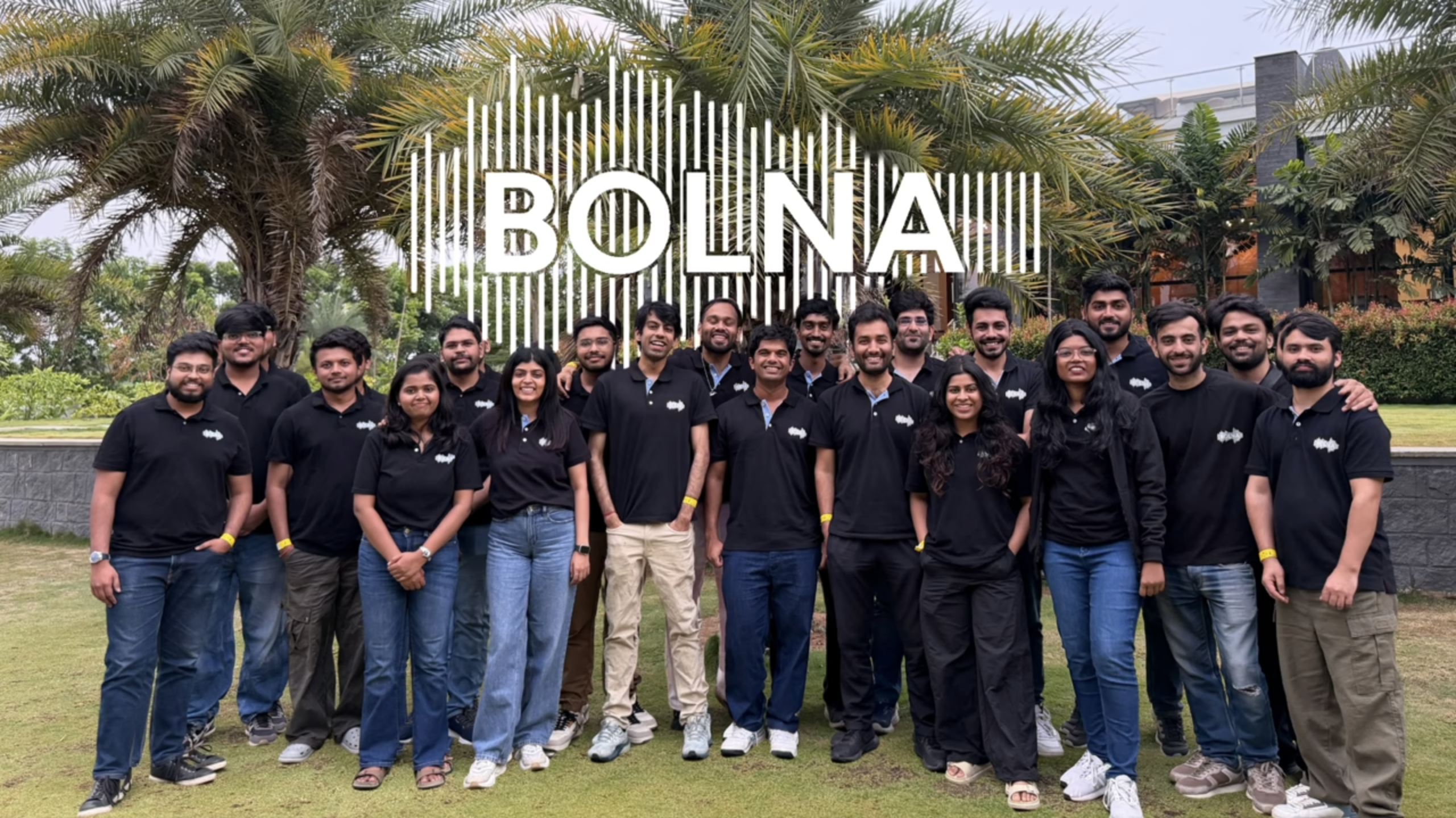 A large group of people posing together outdoors in front of palm trees and greenery. Most of them are wearing matching black polo shirts with a small white logo on the chest. Superimposed over the top of the image is the word 'BOLNA' in large, white, capitalized letters, backed by a white graphic resembling an audio waveform