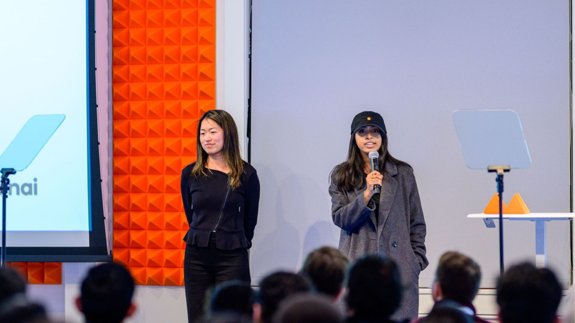 Two women presenting on a stage to an audience. The woman on the right is speaking into a handheld microphone, wearing a long grey coat and a black baseball cap. Her co-presenter stands to her left wearing a black long-sleeved top. Behind them is a presentation screen showing a partial logo and a wall decorated with bright orange acoustic foam panels